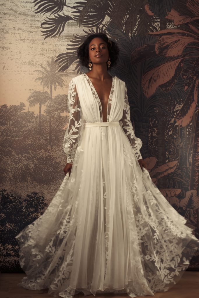 Femme debout en robe de mariée sur mesure Maison Jasmée en tulle brodé de fleurs décolleté devant avec des manches longues