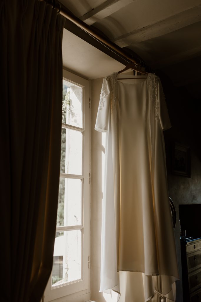 Robe de mariée sur mesure Maison Jasmée en crêpe et boléro suspendu à un cintre devant une fenêtre