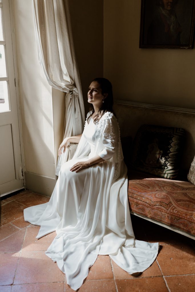 Femme assise sur un divan en robe de mariée sur mesure Maison Jasmée en crêpe avec son boléro