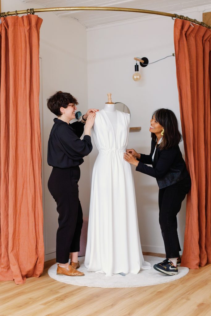 Deux femmes, les créatrices de la maison Jasmée rigolent en ajustant une robe sur un mannequin.