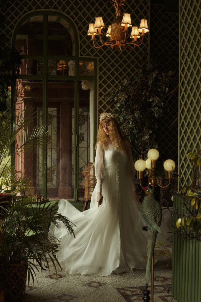 Femme debout portant un ensemble de mariée composée d'une élégante robe à bretelle recouvert d'un voluptueux manteau en organza. Son visage de profil est tourné vers la gauche est son regard se dirige vers le sol.