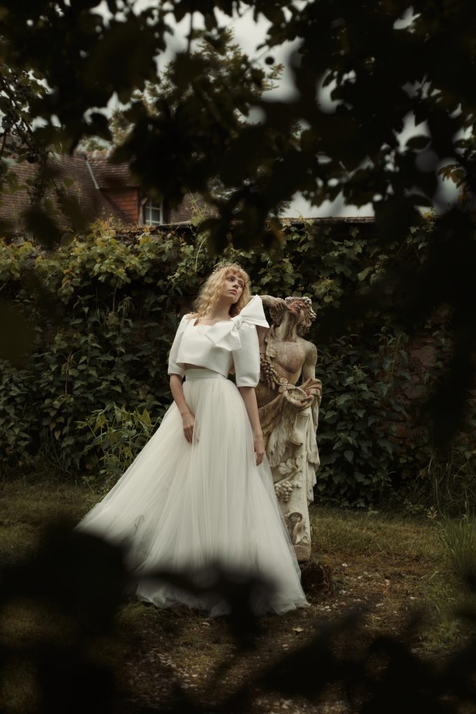 Femme debout de profil adossée à statue en pierre. Elle porte un ensemble de mariée sur mesure composé d'un top en mikado de soie et d'une jupe en tulle.