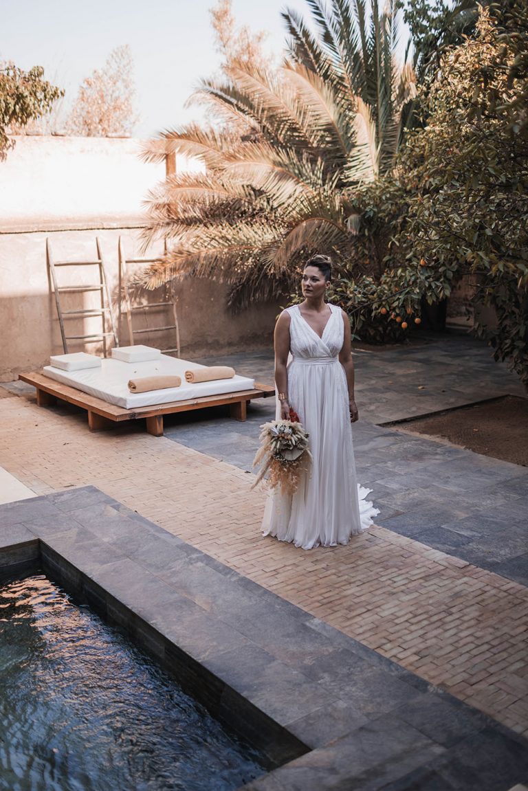 Femme en robe de mariée Maison Jasmée. Elle est au bord d'une piscine et regarde sur sa gauche.