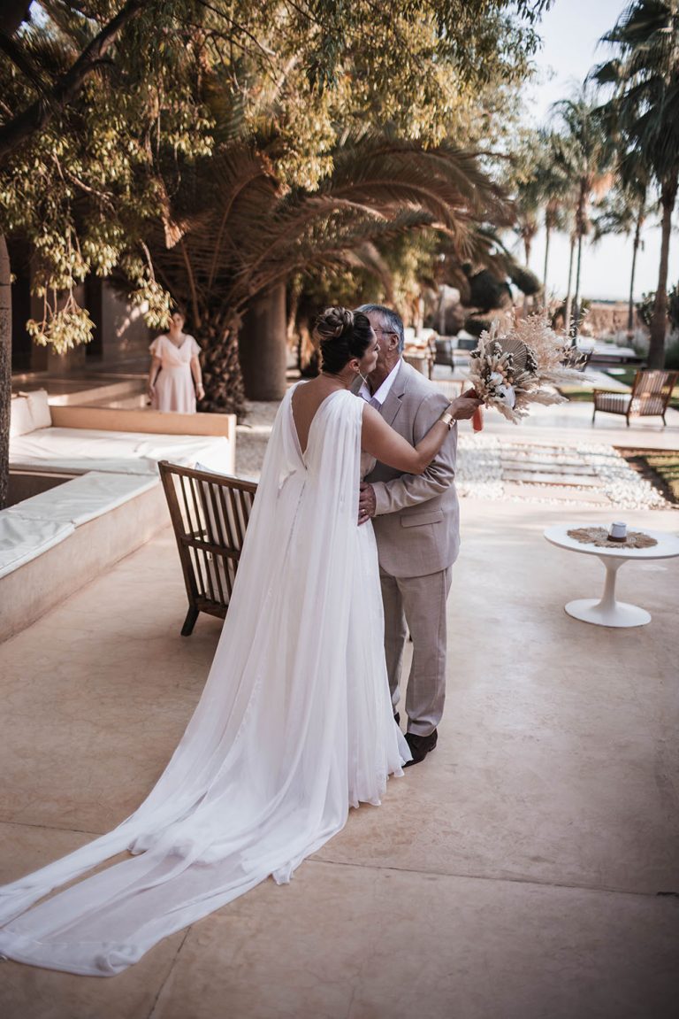 Mariée de dos laissant apparaitre les longs pans de sa robe elle embrasse la joue de son père.