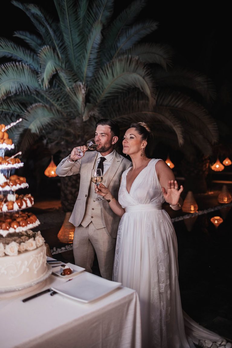 Couple de mariée devant une pièce montée. L'homme bois un verre de vin et la femme tient le sien dans sa main droite.