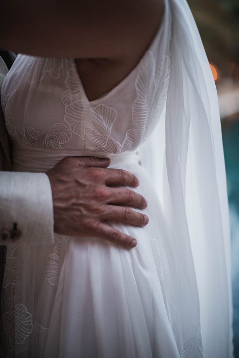 Détail du profil d'une robe de mariée sur mesure Maison Jasmée. Nous voons la main d'un homme posé sur la taille de sa femme.