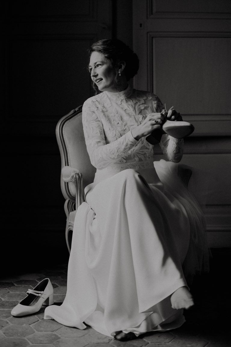 Femme assise en robe de mariée Maison Jasmée s'apprêtant à enfiler sa chaussure.