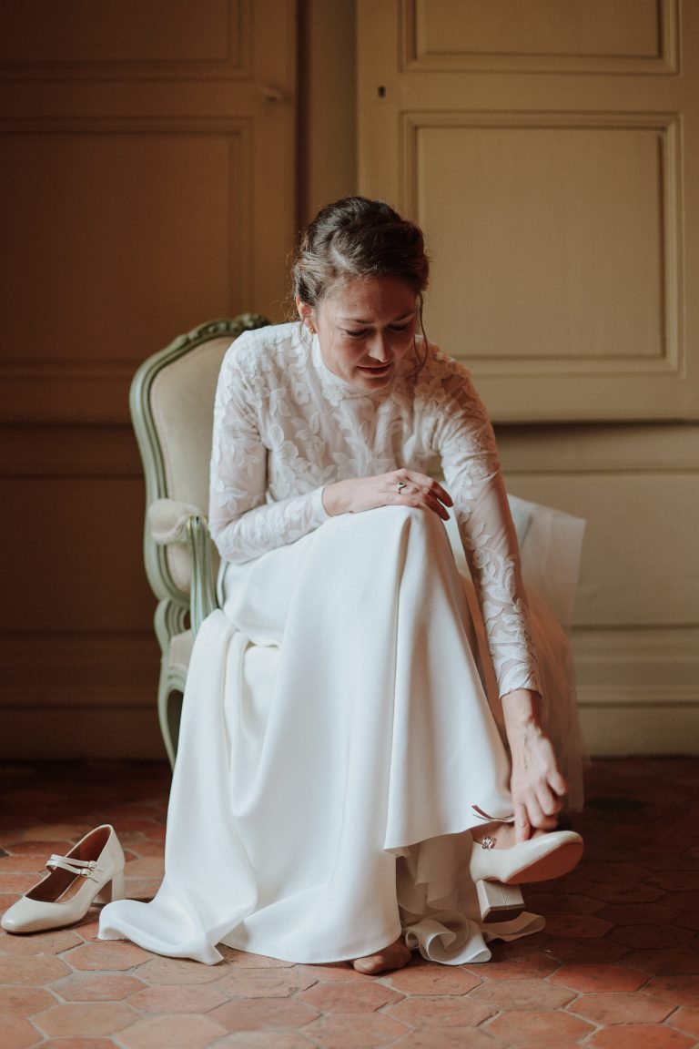 Femme assise en robe de mariée Maison Jasmée attachant ses chaussures à talons.