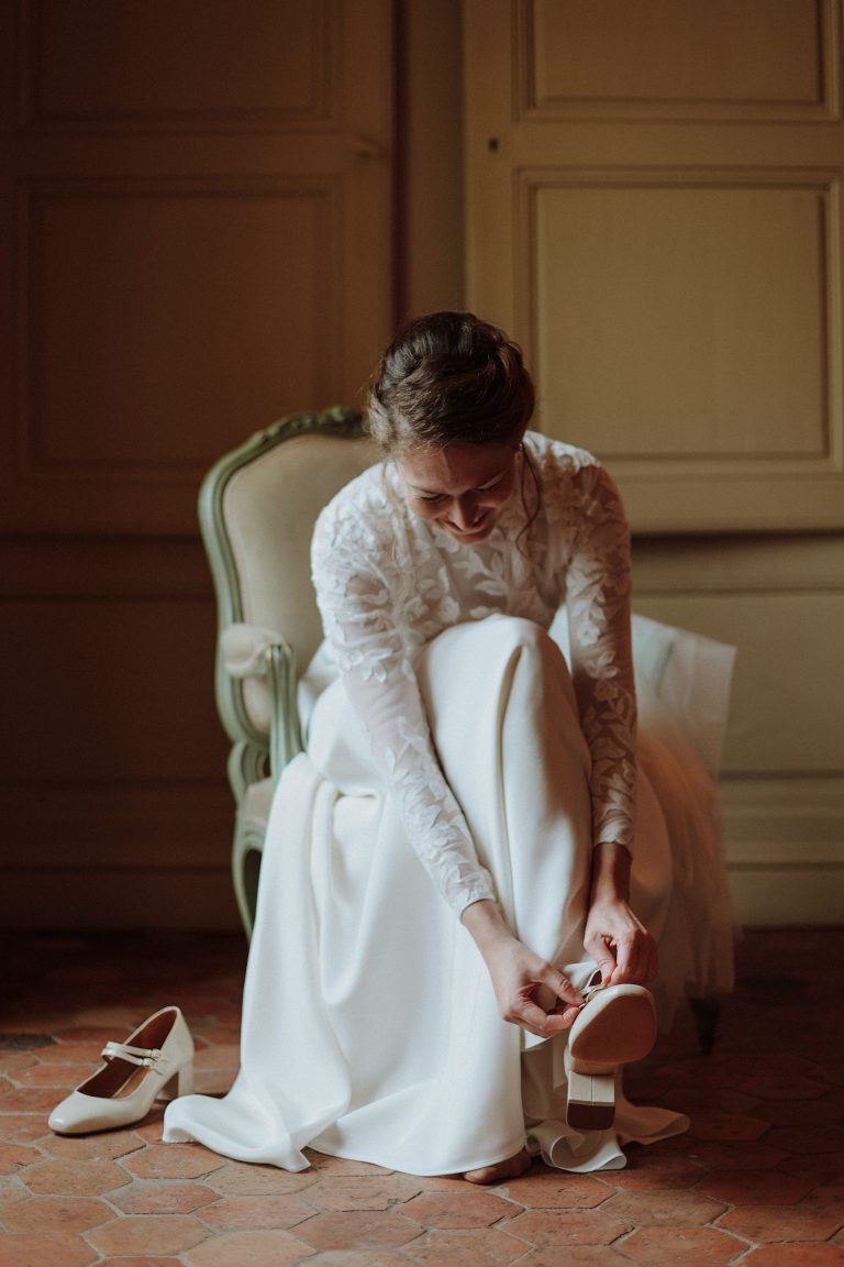 Femme assise en robe de mariée maison jasmée attachant ses chaussures à talons.