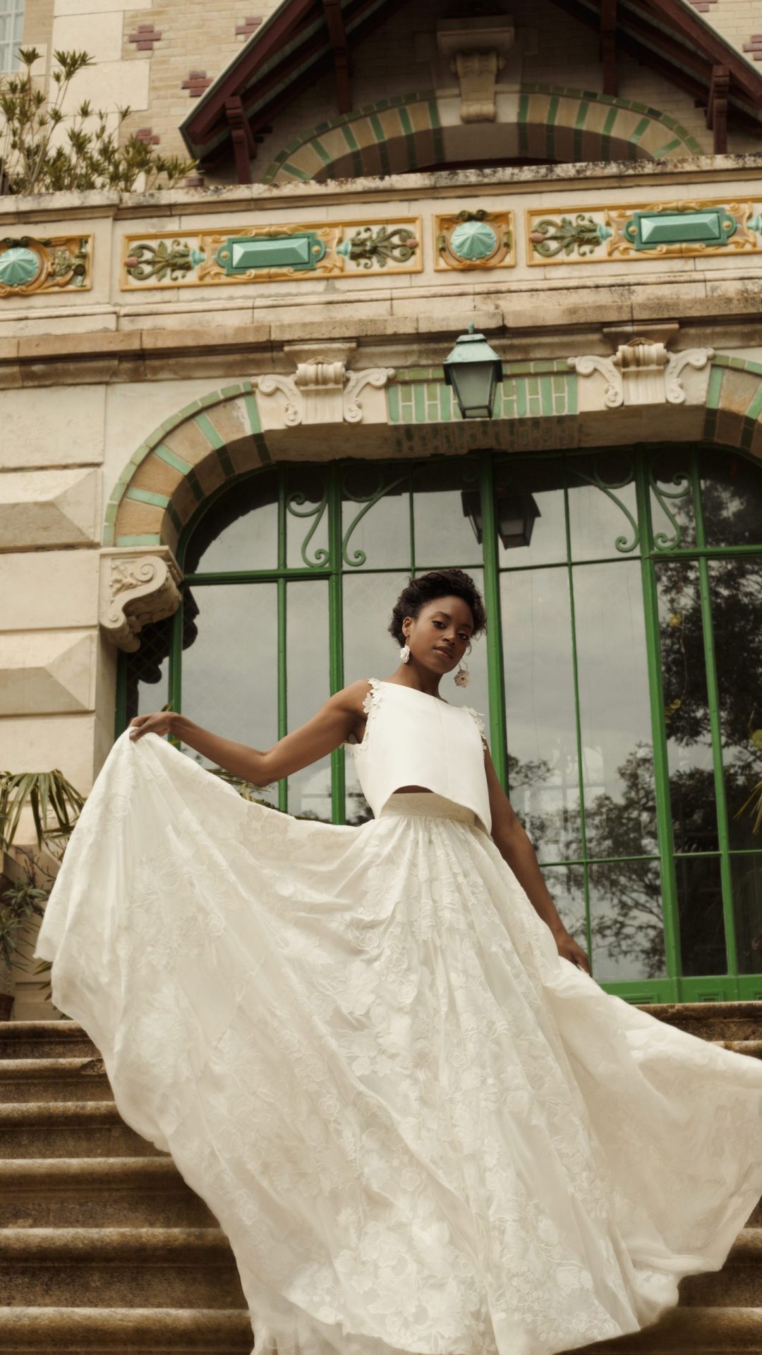 Femme debout de face tenant dans ses mains une voluptueuse jupe en crèpe georgette de soie brodé. Elle porte également un top en drap de soie brodé de fleurs de guipure et de perles.