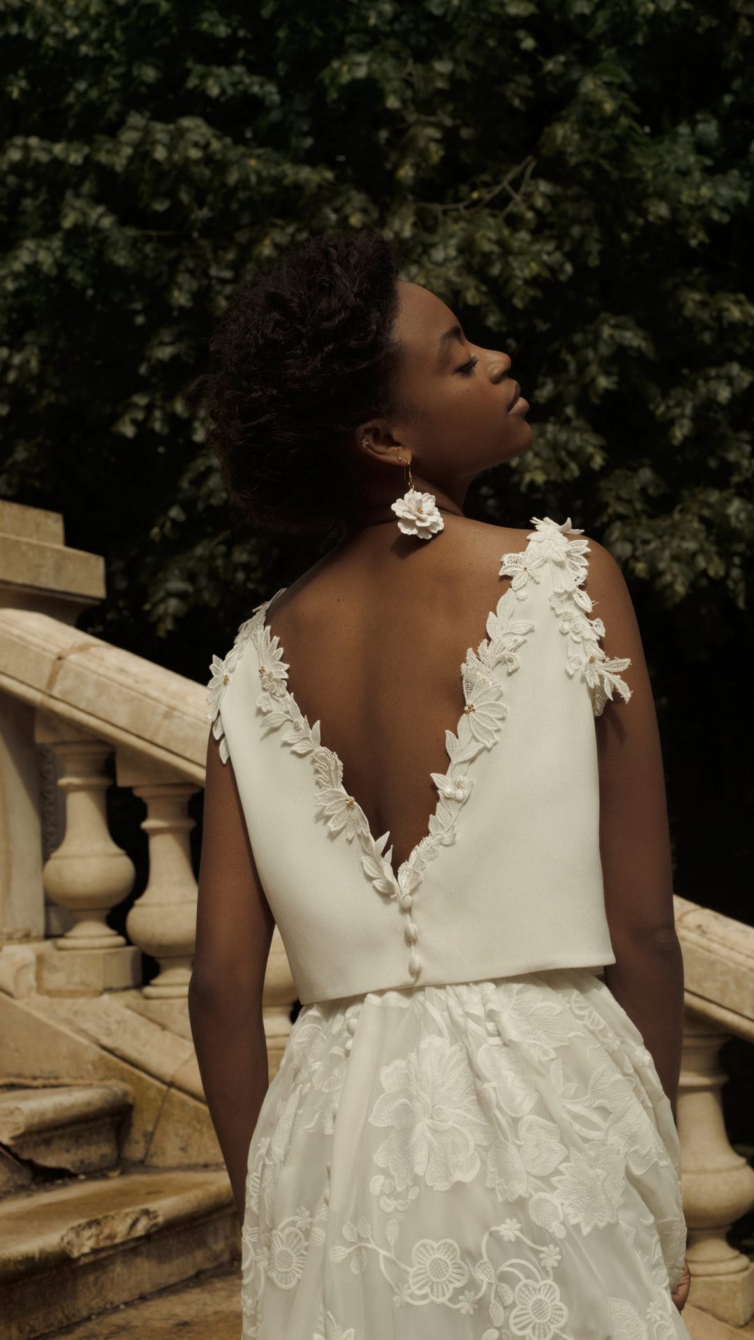 Femme debout de dos. C'est un plan de détail du top qu'elle porte en drap de soie dont le décolleté dorsale est brodé de fleurs de guipure et de perle.