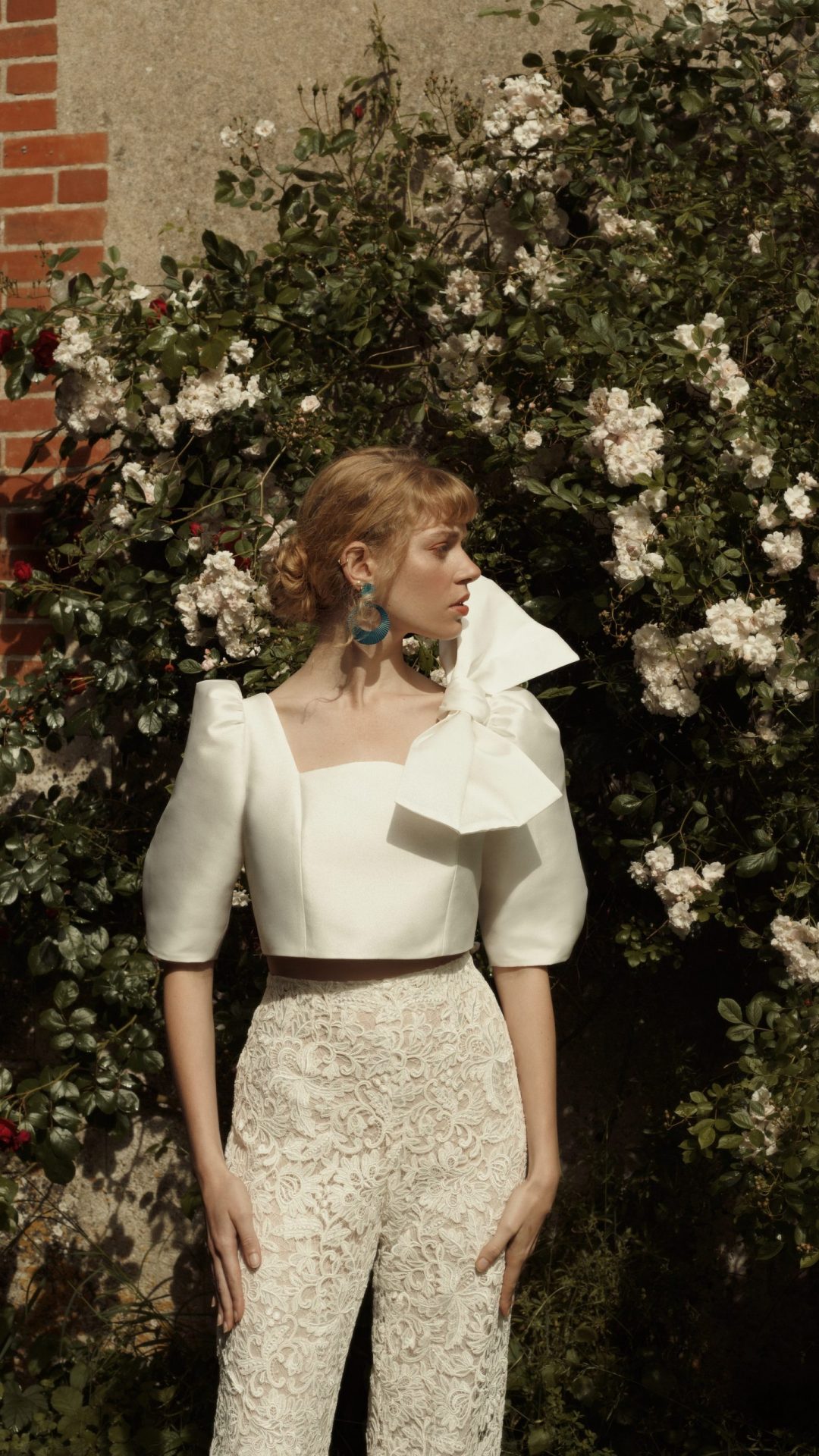 Femme debout le visage de profil. Elle se tient devant des fleurs et porte un ensemble de mariée nommé Soho composé d'un top en mikado de soie décoré d'un noeud papillon XL et d'un pantalon en guipure.