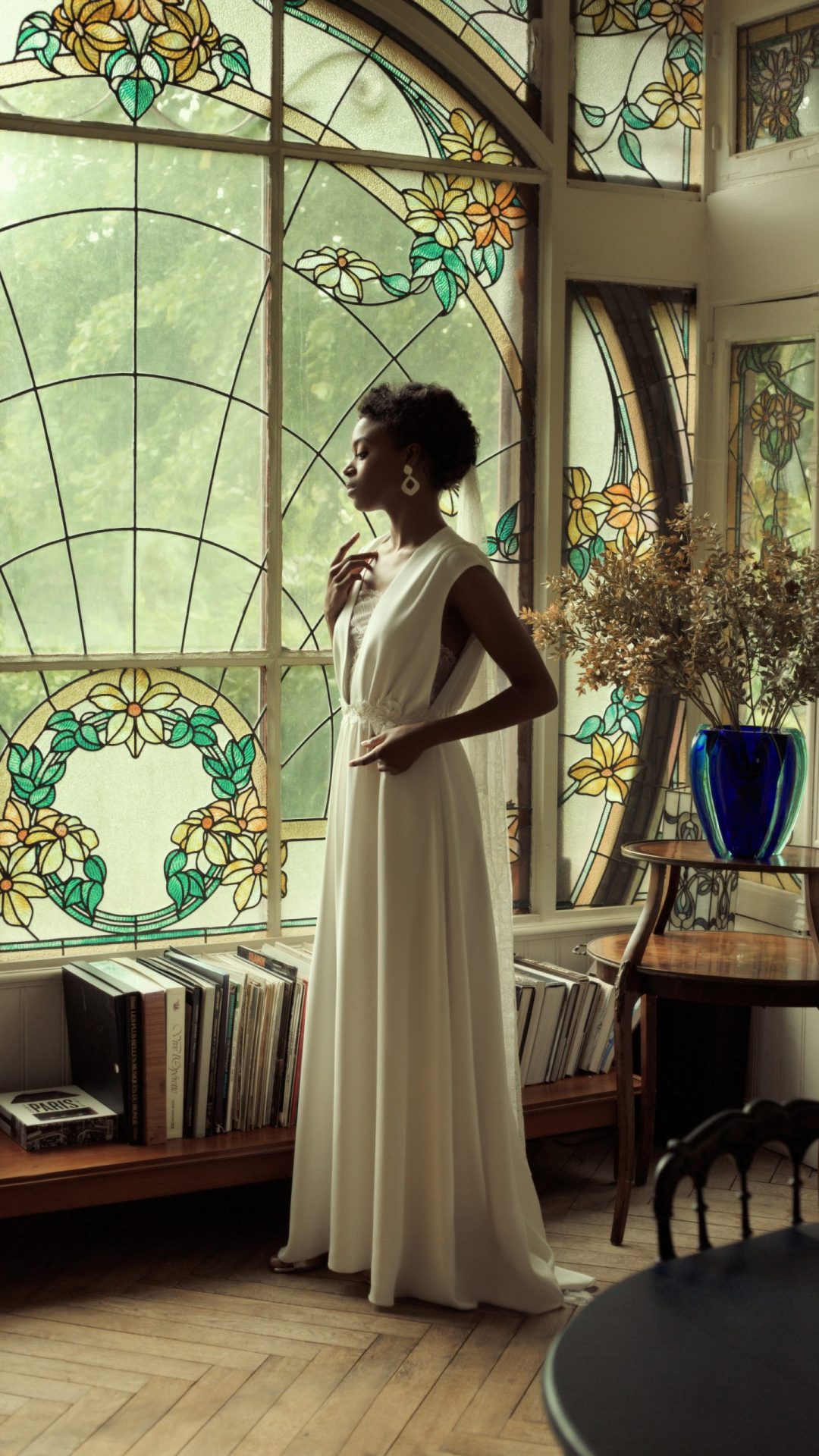Femme debout, regardant en direction d'une fenêtre. Elle porte une robe de mariée en drap de soie aux lignes sobres.