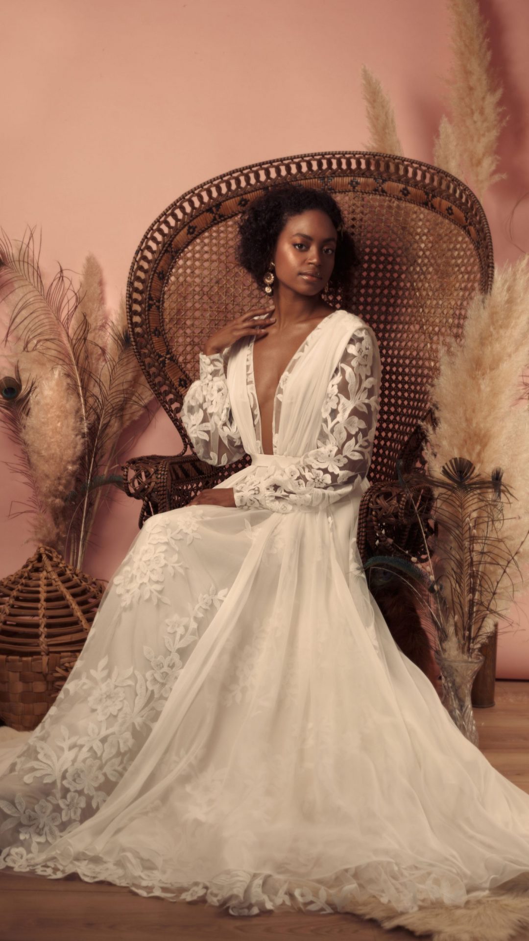 Femme assise sur un fauteuil en rotin en robe Maison Jasmée en tulle brodé de fleurs avec des manches longues et un décolleté en V devant